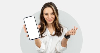 Bir kadın, bir elinde boş bir ekranla akıllı bir telefonu tutarken diğer eliyle el kol hareketi yapıyor. O, beyaz bir gömlek giydi ve parlak bir gülümsemesi var. Arka plan sade..