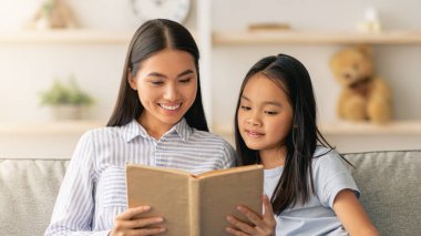 Aile eğlencesi konsepti. Genç Asyalı anne ve kızı birlikte kitap okuyor, kanepede oturuyor, boş yer var. Mutlu anne ve çocuk evde boş vakit geçirmekten zevk alıyor.