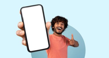 Kıvırcık saçlı genç bir adam boş bir akıllı telefon ekranını heyecanla gösteriyor. Gülümsüyor ve açık mavi bir arkaplanda baş parmak işareti yapıyor. Neşeli ve çekici bir hava var..