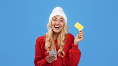 Siber alışveriş konsepti. Altın kredi kartını elinde tutan neşeli mutlu kadın akıllı telefon kullanıyor, beyaz kış şapkası takıyor mavi stüdyo arka planında alışveriş torbalarıyla yerde oturuyor.