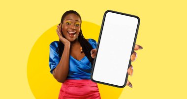 Neşeli bir kadın, elinde boş bir telefonla parlak sarı bir arka plana karşı poz veriyor. Renkli kıyafeti ve neşeli ifadesi hayat dolu bir atmosfer yaratıyor..