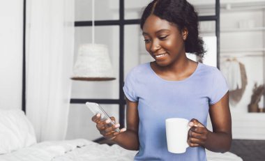 Neşeli siyah kadın interneti tarıyor ve evde modern yatak odasında yatağında oturmuş kahve içiyor. Teknoloji ve Malzemeler, Mobil Teklif, Yeni Uygulama Konsepti