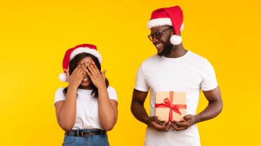 Noel Baba Şapkalı Afro-Amerikalı Adam, Sarı Stüdyo 'nun arka planında gözlerini kapayarak Noel Hediyesi Beklerken Sürpriz Kadın Tutuyor. Mutlu yıllar, Xmas Sunar Teslimat Konsepti