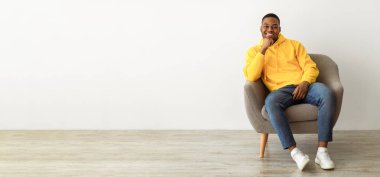 Başarılı Siyahi Milenyum Adamı Kameraya Gülümseyen Chin 'e Dokunan Kol Sandalyesinde oturan Sarı Kapüşonlu Kapüşonlu Adam. Afro-Amerikalı adam evde rahatlama pozu veriyor. Tam Uzunluk