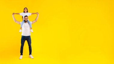 Sarı Stüdyo Arkaplanı Üzerine Boynunda Oturan Mutlu Arap Kızı. Baba, çocuğu kucağında tutuyor ve kameraya gülümsüyor. Birlikte eğlenceli pozlar veriyor.