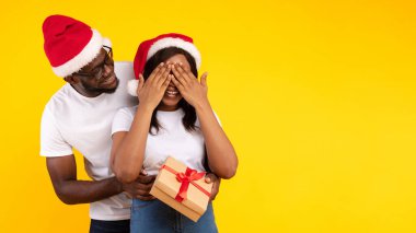 Noel hediyesi. Stüdyoda sarı arka planda duran Noel Baba Şapkalarını giyerken Xmas Hediyesiyle Kız Arkadaşı Şaşırtan Afrikalı Amerikalı Adam. X-Mas ve Yeni Yıl Tatil Sezonu