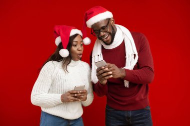 Heyecanlı Afrikalı Amerikalı Adam Kız Arkadaşına Akıllı Telefon Gösteriyor Telefon Duruşu İçin Yeni Harika Cep Telefonu Uygulaması Öneriyor Sarı Arkaplan Üzerinde Noel Baba Şapkaları Giyiyor, Stüdyo Çekimi. İnsanlar ve Gadget