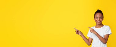 Beyaz tişörtlü, gülümseyen genç siyah kız sarı arka planda, panorama 'da izole bir şekilde boş alanı işaret ediyor. Pozitif duygular, harika bir teklif, reklamcılık ve liseli kızdan tavsiye.