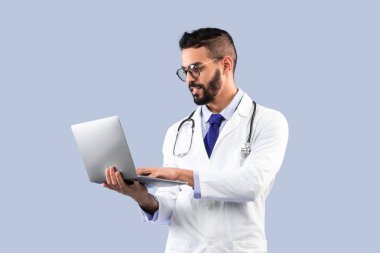 Mavi Stüdyo Arkaplanının Üzerinde Duran Dizüstü Bilgisayarı kullanan Arap Erkek Doktor. Doktor Adam, beyaz üniformalı, reklamcı, bilgisayarlı tıbbi web sitesi pozu veriyor. Dikey Çekim