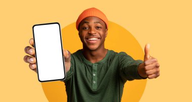 Genç bir adam parlak sarı bir arkaplana karşı poz veriyor, boş bir ekranla akıllı telefonu gösteriyor. Geniş bir gülümseme sergiliyor ve coşkuyla olumlu bir atmosfer yaratıyor..