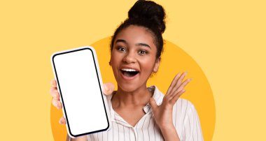 Genç bir kadın, canlı sarı bir arkaplan önünde akıllı bir telefonu tutarken geniş bir şekilde gülümser. Gösterdiği cihazdan duyduğu neşeyi ifade ederken heyecanı çok açık..