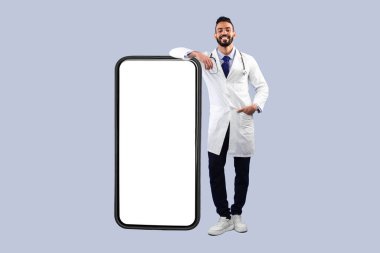 Orta Doğulu erkek doktor, mavi arka planda duran boş ekran pozuyla büyük akıllı telefona yaslanıyor. Profesyonel Hekim Reklamcılığı Tıbbi Mobil Uygulaması. Model, Meydan