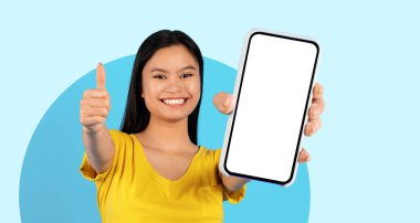 Uzun siyah saçlı genç bir kadın bir elinde telefonu tutarken geniş bir şekilde gülümsüyor ve diğer eliyle başparmağını kaldırıyor. O sarı bir gömlek giyiyor ve arka planı yumuşak bir mavi..