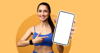 Mavi aktif giysili gülümseyen bir kadın onu işaret ederken boş bir akıllı telefon ekranı tutuyor. Parlak turuncu bir arka plana karşı. Terfi için canlı bir atmosfer öneriyor..