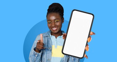 Neşeli genç bir kadın, parlak mavi bir arka plana karşı duruyor. Elinde boş bir akıllı telefonu tutarken izleyiciye başparmak işareti yapıyor. Yüzü neşeli ve çekici..