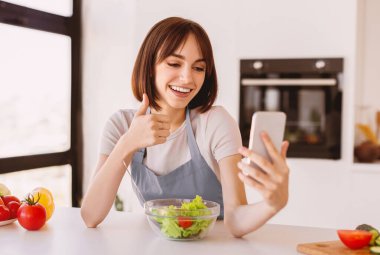 Sağlıklı yiyeceklerle ilgili video blogu. Taze salatalı mutlu bayan akıllı telefondan video görüşmesi yapıyor ve baş parmağını kaldırıyor, mutfakta duruyor, yeni vlog kaydediyor.