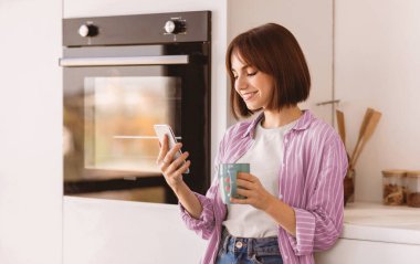 Sosyal ağlar ve sabahları sohbet. Mutlu genç bayan kahve içiyor ve akıllı telefon kullanıyor, modern mutfağın içinde duruyor. Heyecanlı bir kadın arkadaşlarıyla mesajlaşıyor ve sıcak içkinin tadını çıkarıyor.