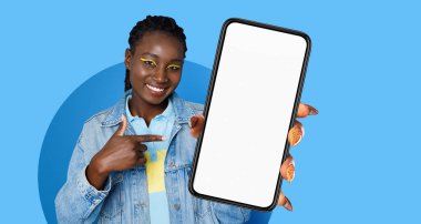 Bir kadın parlak mavi bir arka planda boş bir akıllı telefon ekranına işaret ederken gülümsüyor. Şık bir makyaj ve günlük kıyafetler giyer. Seyirciler için davetkar bir ortam yaratır..