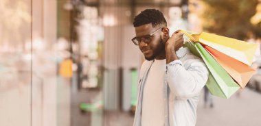 Gözlüklü yakışıklı zencinin portresi moda kıyafetlerine bakıyor ya da alışveriş merkezinin vitrininde yeni ürünlere bakıyor, alışveriş merkezinin yakınında çantalarını tutuyor. Hediyeler, Satış ve İndirim, Perakende, Satın Alma Konsepti