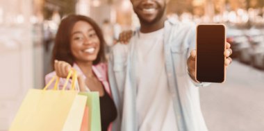 Çevrimiçi Mağaza Uygulaması. Mutlu siyah çift kameraya yakın boş ekranlı akıllı telefonları gösteriyor ve ellerinde çantalar, dışarıda duran neşeli insanlar, Mock Up, bedava fotokopi alanı.