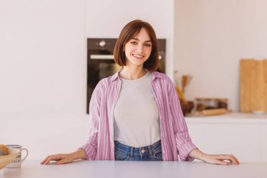 Masada poz veren, mutfakta duran, hafta sonu evde dinlenirken kameraya bakıp gülümseyen mutlu genç bir ev hanımı.