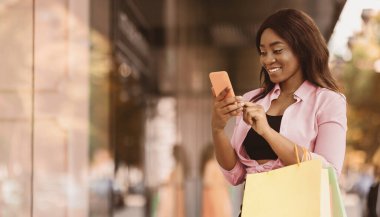İnternetten alışveriş. Gülümseyen siyah kadın cep telefonunu kullanıyor, alışveriş torbalarını tutuyor, alışveriş merkezinin penceresinin yanında duruyor, kıyafet ve hediyeler seçiyor, fotoğraf çekiyor, boş yer arıyor. Özel Teklif