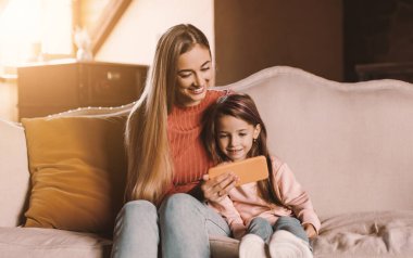 Havalı Gadget ve Uygulama. Oturma odasında kanepede oturan, cep telefonu kullanan, gülümseyen genç bir kadın ve küçük bir kızın portresi. Ailece internette gezinmek, birlikte çizgi film izlemek, bedava kopya alanı