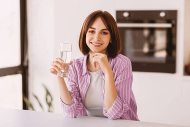 Camdan su içen genç bir bayanın portresi. Mutfakta oturup kameraya gülümsüyor. Mutlu kadın masada oturuyor ve sağlıklı bir sıvı tutuyor. Vücut sulanmasına özen gösteriyor.
