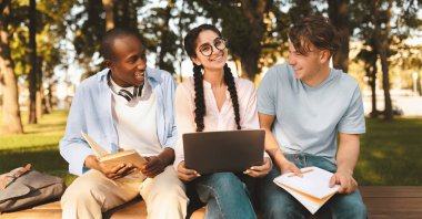 Farklı öğrenci arkadaşları üniversite kampüsünde bankta oturarak öğreniyor. Sınıf arkadaşları laptopla dinleniyor ve parkta bir şeyler tartışıyorlar.