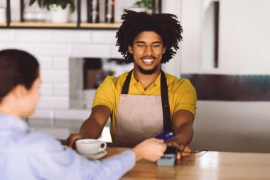 Tezgahta önlüklü, sırıtan kıvırcık, çekici, genç, zenci bir barmen kadın müşteriye kafenin içinde kredi kartı veriyor. Düzen, finans, iş ve covid-19 'a bağlı küçük işletmeler
