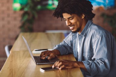 Kumanda Ofisi. Genç Afro-Amerikan iş adamı, kafede dizüstü bilgisayarla çalışırken akıllı telefon kullanıyor, siyah milenyum girişimcisi gülümsüyor müşterilerle iletişim kurmak için cep telefonu kullanıyor, uzaylıları kopyalıyor.