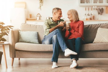 Mutlu son sınıf çifti ellerinde sıcak içecekle kahve fincanları tutuyorlar ve evdeki koltukta oturup birbirlerine gülümsüyorlar. Haftasonu. Emeklilik Yaşamı ve Mutlu Evlilik Konsepti