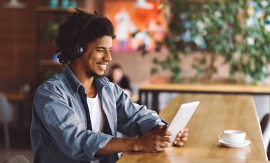 Kablosuz kulaklıklı mutlu, kıvırcık, genç siyah erkek tablete bak ve kafenin içinde müzik dinle, profil. Ses uygulaması, duygular ve yüz ifadesi, komik video, boş zamanlardaki sosyal medya