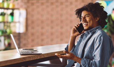Gülümseyen milenyum Afro-Amerikan kıvırcık adamı akıllı telefonuyla konuşuyor, masada dizüstü bilgisayarla oturuyor, kafenin içinde not defteri var. Bağımsız yüklenici için uzaktan çalışma, birlikte çalışma alanındaki istemciye çağrı