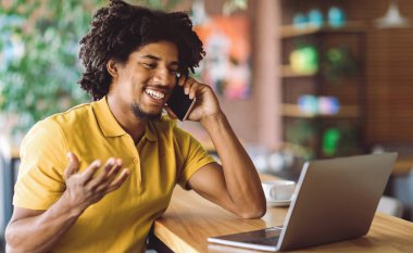 Uzaktan kumanda. Cafe 'de dizüstü bilgisayar kullanan ve cep telefonuyla konuşan neşeli siyahi genç adam, Milenyum Afrikalı Amerikalı Serbest Çalışan ve Cep Telefonu Konuşmasının tadını çıkaran,