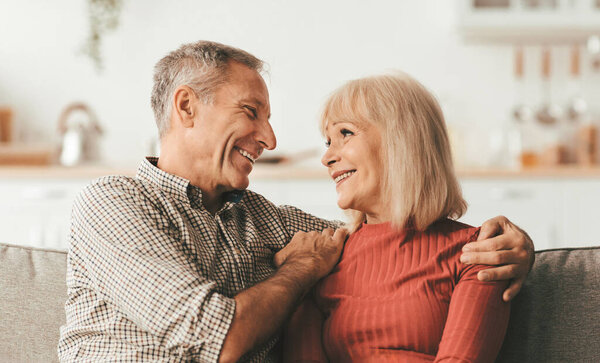 Веселый зрелый муж обнимает счастливую жену, улыбающуюся ей, сидя дома на диване. Уход в отставку и семейный образ жизни. Романтические отношения в пожилом возрасте