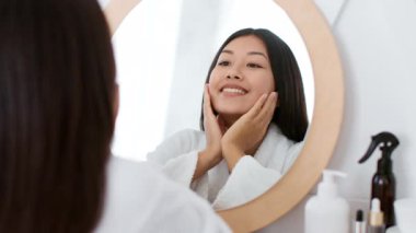 Bu genç Asyalı kadın evinde kendini şımartıyor. Aynanın karşısına geçmiş gülümseyip güzellik rutininin tadını çıkarırken cilt ürünlerini uyguluyor. Doğal ışık rahatlatıcı atmosferi arttırır..