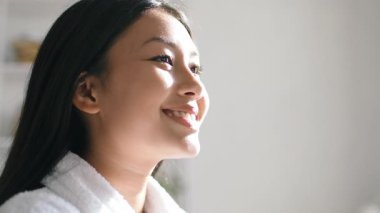 Genç bir Asyalı kadın rahat bir evde kendine zaman ayırırken gülümsüyor. Yumuşak doğal ışık, rahatlama ve şımartma anlarını kucaklarken cilt bakımını arttırır..