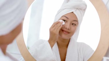 Huzurlu bir ev ortamında, genç bir Asyalı kadın dairesel aynaya bakarken cilt bakım ürünlerini pamukla uyguluyor. Üzerinde sabahlık ve havlu var. Rahatlama ve kendine bakım yayıyor..