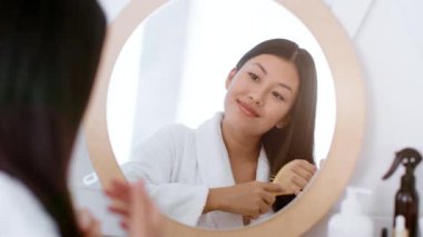 Genç bir Asyalı kadın saçını tararken evde rutinini gerçekleştiriyor. Aynadaki yansımasına gülümsüyor, rahatlatıcı bir kişisel bakım anının tadını çıkarıyor..