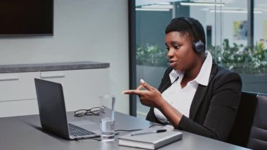 Profesyonel kıyafetli genç bir Afrikalı Amerikalı kadın çağdaş bir ofis ortamında bir video konferansı düzenliyor. Gösterişli bir masada otururken odaklanmış ve meşgul..