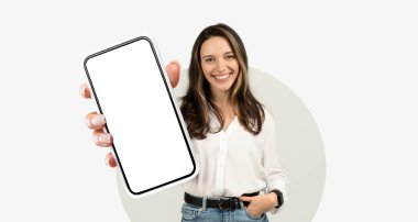 Neşeli bir kadın, akıllı telefonuna boş bir ekran sunuyor. Pozitif tavrını gösteriyor. Hafif bir arka planda duruyor, günlük giysiler içinde..