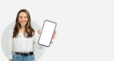 Genç bir kadın parlak, modern bir ortamda duruyor, bir elinde akıllı telefonu tutarken gülümsüyor. Arka plan basit, dikkatini onun neşeli tavırlarına odaklıyor..