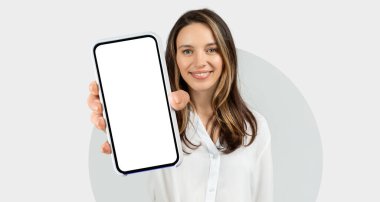 Genç bir kadın stüdyo ortamında akıllı bir telefonu tutarken parlak bir şekilde gülümsüyor. Uzun saçları var ve beyaz bir gömlek giyiyor. Arka plan basit ve temiz..