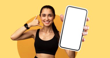 Genç bir kadın akıllı telefon tutarken gülümsüyor ve jest yapıyor. Günlük atletik kıyafetler giyiyor ve parlak sarı arka plan sahneye enerji katıyor..