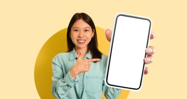 Genç bir kadın elinde tuttuğu boş bir akıllı telefona gülümseyen ve işaret eden parlak sarı bir arka plan karşısında duruyor. Ayar, pozitif ve canlı bir atmosfer taşıyor..