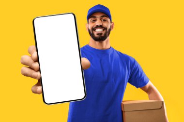 Boş ekranlı ve karton kutulu cep telefonunu gösteren mutlu Arap kurye teslimat hizmeti veren, sarı arka planda poz veren adam. Stüdyo Çekimi, Model