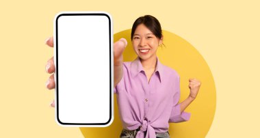 Parlak sarı bir ortamda, genç bir kadın gülümser ve boş bir akıllı telefon gösterir. Lavanta bir bluz giyiyor ve neşeli bir pozla kendine güveni var..