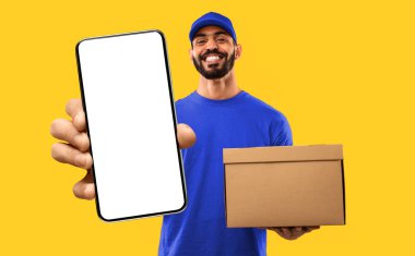 Teslimat işi. Sarı Stüdyo Arkaplanı Üzerinde Para ve Paket Kutusu Poz Veren Kırmızı Üniformalı Neşeli Arap Kurye. Ulaştırma ve Nakliye Servisi