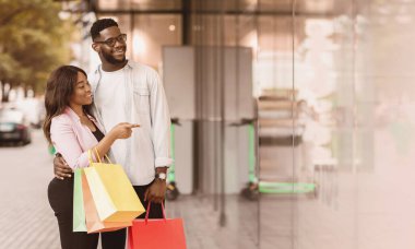 Alışverişkolik kavram. Afro-Amerikalı genç bir kadının tam boy profili. Parlak alışveriş torbaları taşıyan adama sarılıyor. Vitrinlere mankenlerle bakıyor.